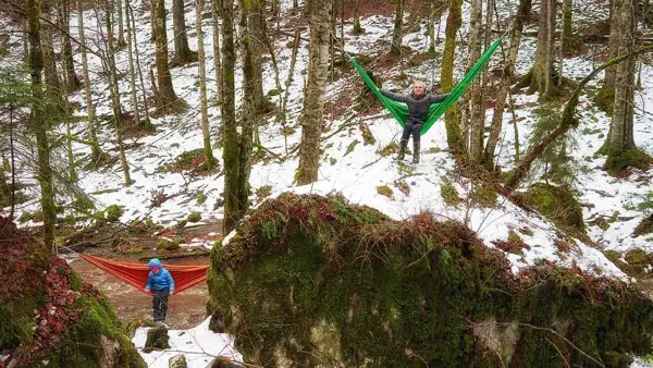 To personer i hver sin hengekøye mellom trær i delvis snødekket skog
