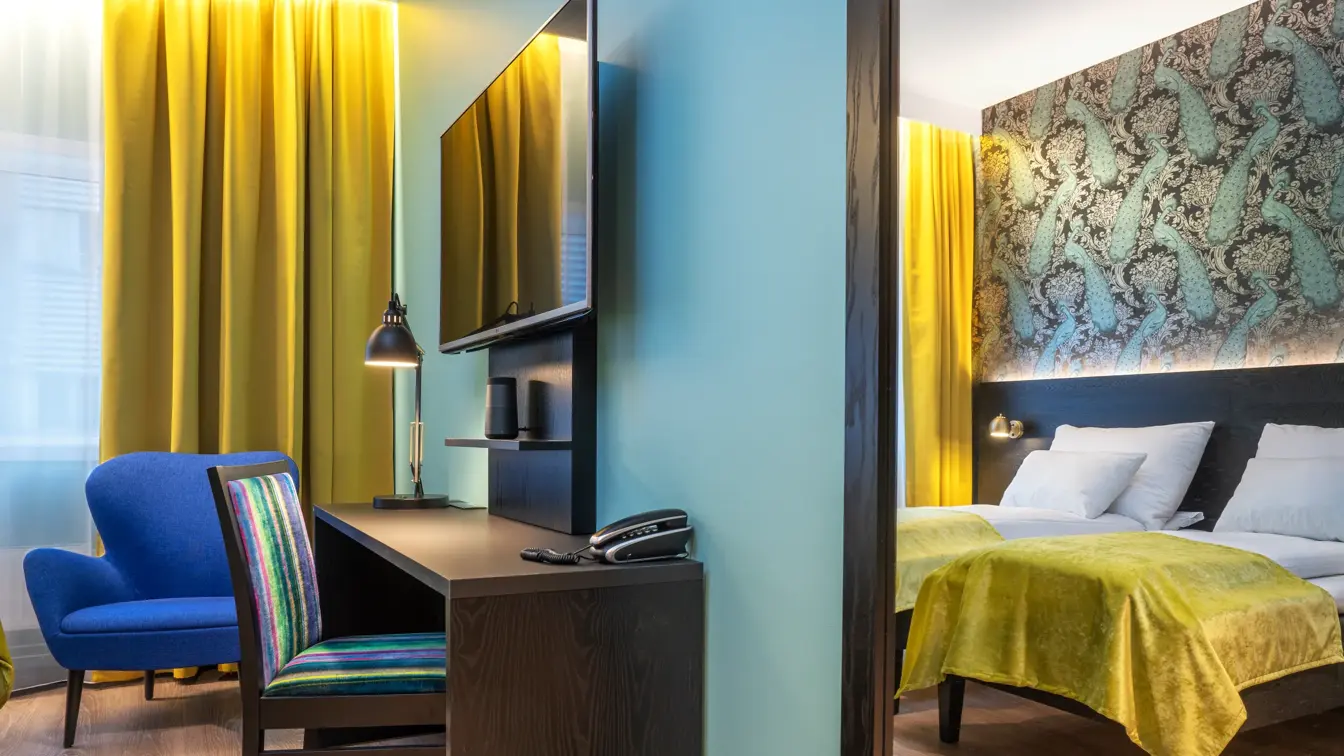 Gule gardiner, vindu fra gulv til tak, to enkel senger, tapetvegg me dmønster, skrivebord, TV, blå stol, skrivebordstol, parkettgulv. Speil på veggen. 