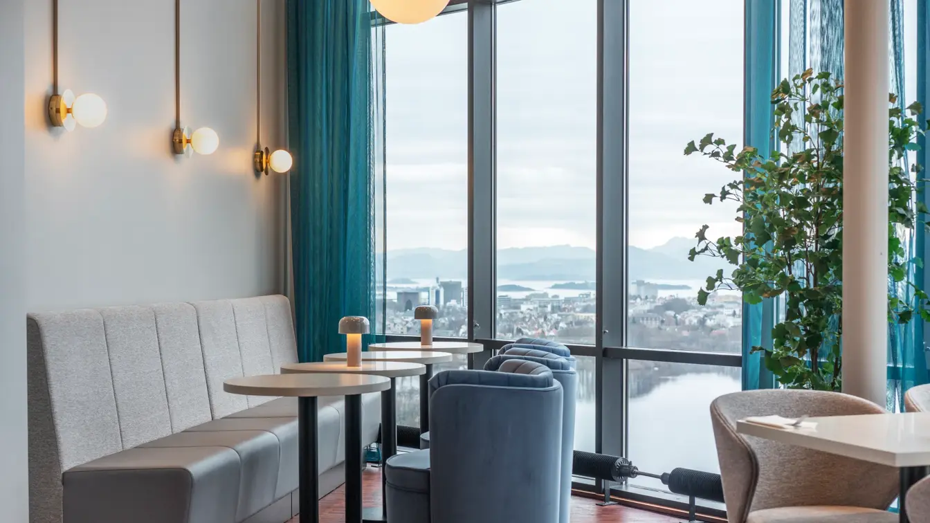 Grå sofa inntilvegg, runde bord, blå stoler, gulv-til-tak-vinduer i restaurant.