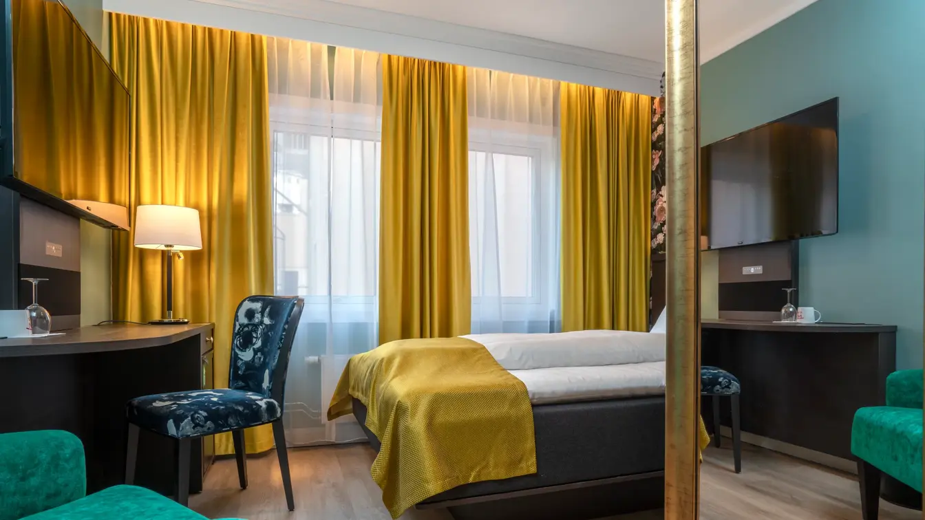 Dobbeltseng, vindu, gule gardiner, skrivebord, grønn lenestol, TV, blå stol, veggspeil, grønne vegger, parkettgulv