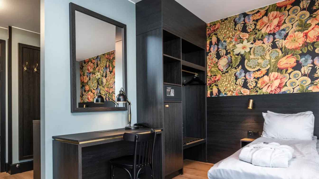 Business rom med seng, åpen garderobe, skrivebord, stol, speil og fargerik tapet.