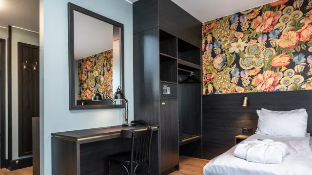 Business rom med seng, åpen garderobe, skrivebord, stol, speil og fargerik tapet.