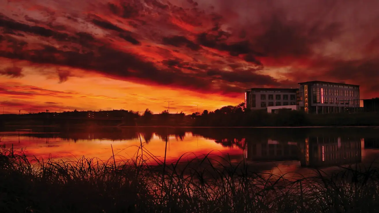 Fasadebilde av Thon Hotel Arena i solnedgang. I forgrunnen ses Nitelva.