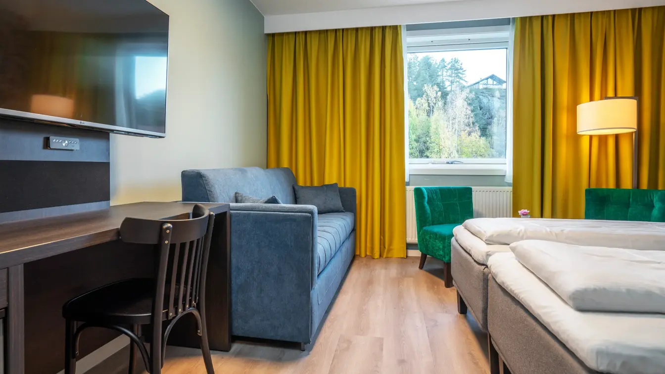 Familierom med twin senger, blå sofa, skrivebord med stol, grønne lenestoler og gule gardiner.