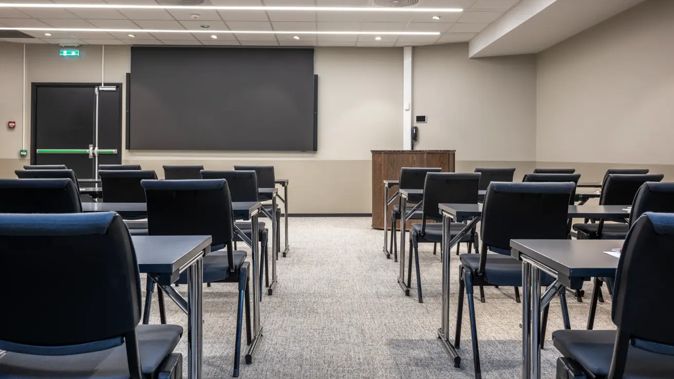 Nærbilde av møterom med klasseroms oppsett sett bakfra i lokalet. Stor skjerm foran sammen med talerstol.