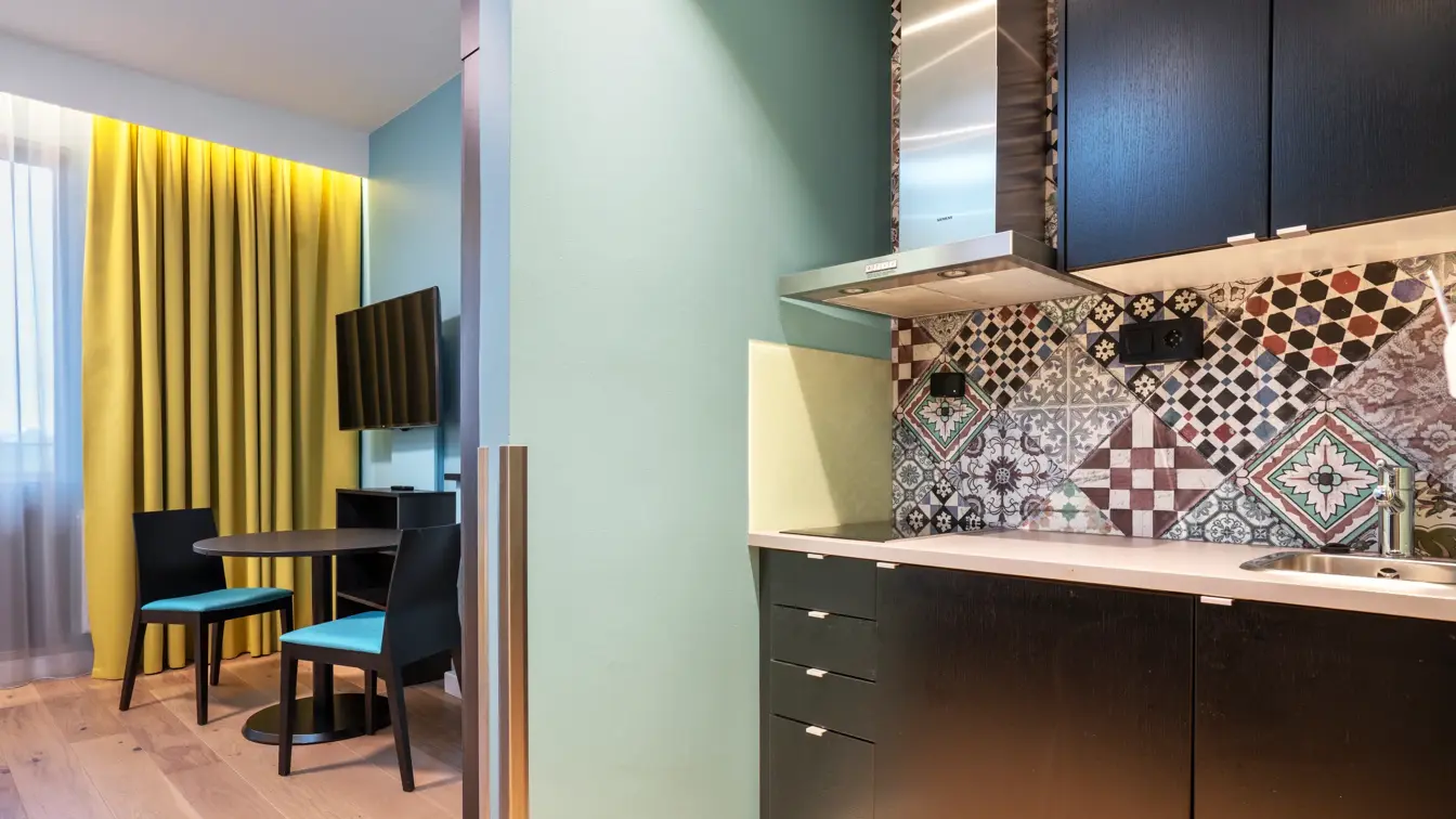 Parkettgulv, kjøkken, blå vegger, gule gardiner, vindu fra gulv til tak, Tv skjerm, to brune stoler, stuebord.