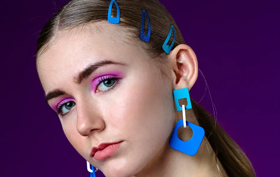 poserende dame foran ensfarget bakgrunn med mønstrete topp, blå øredobber og blå hårspenner