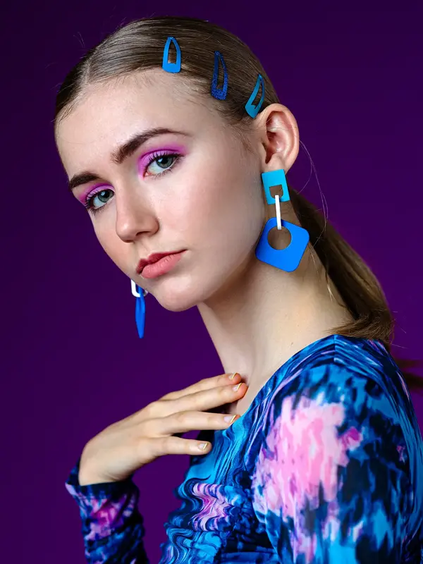 poserende dame foran ensfarget bakgrunn med mønstrete topp, blå øredobber og blå hårspenner