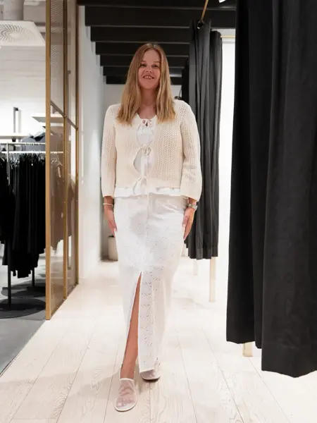 damemodell med hvit topp, hvitt skjørt, beige jakke og hvite sko.