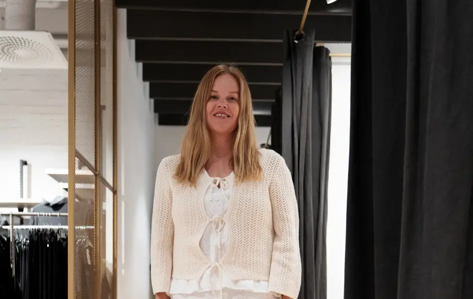 damemodell med hvit topp, hvitt skjørt, beige jakke og hvite sko.