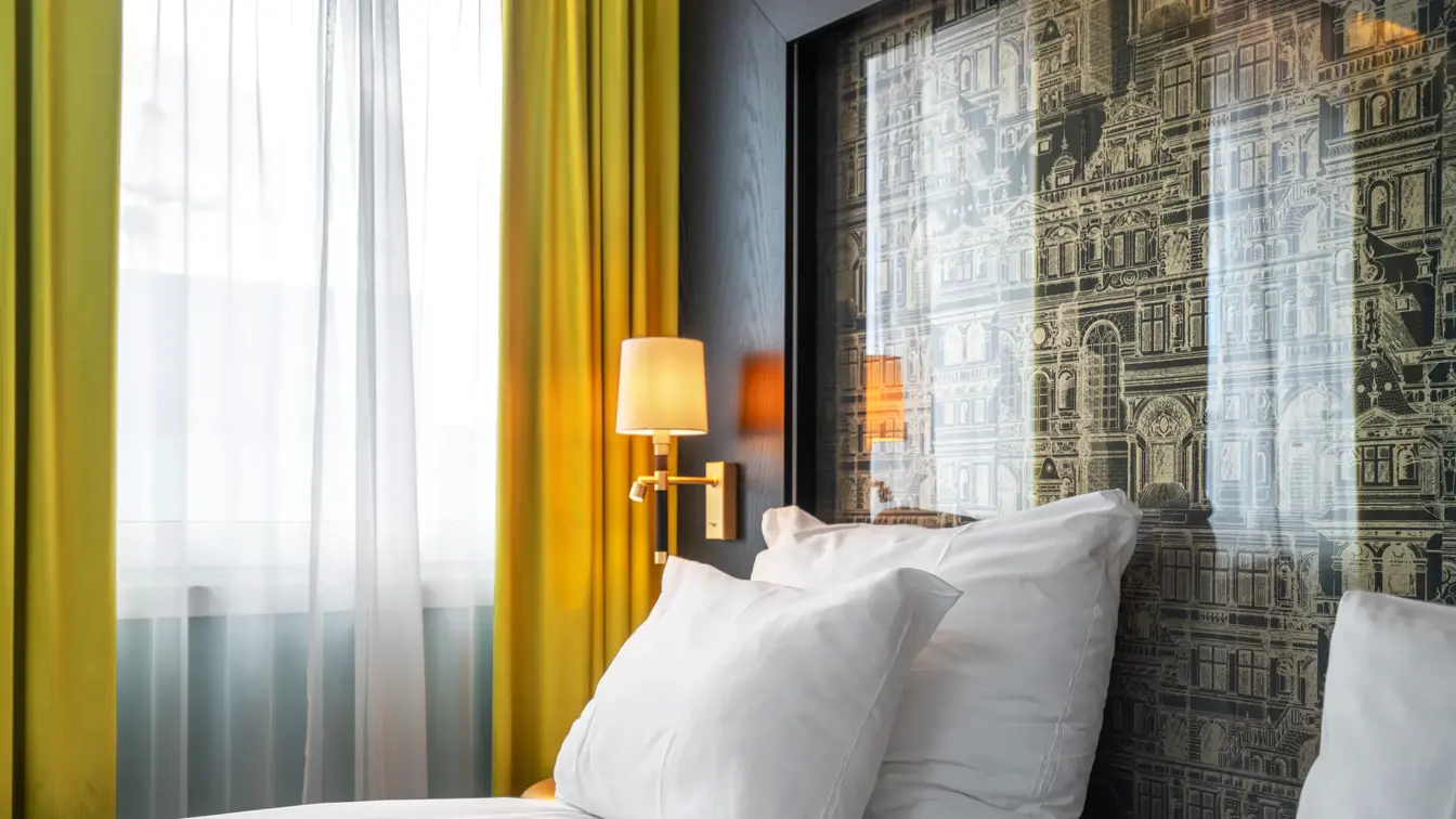 Dobbeltrom med dobbeltseng, hvite puter, gul gardin og tent sengelampe.