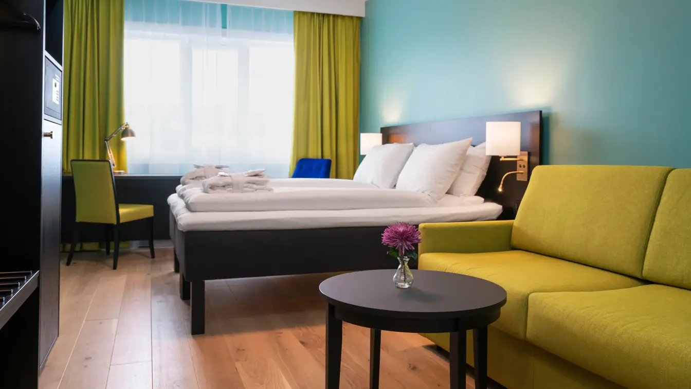 Gul sofa, bord, garderobeskap, dobbeltseng, skrivebord og lenestol på Buisness room på Thon Hotel Europa i Oslo sentrum rett ved Slottsparken
