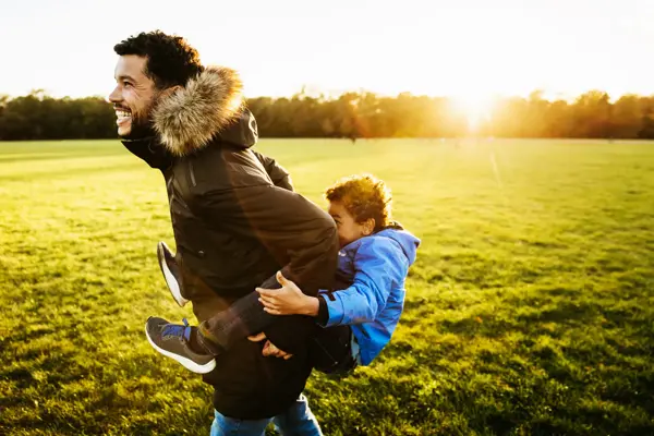 A single dad out with his son at the park, giving him a piggy back ride.