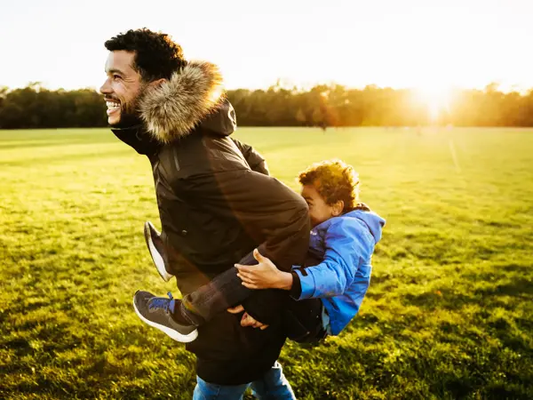 A single dad out with his son at the park, giving him a piggy back ride.