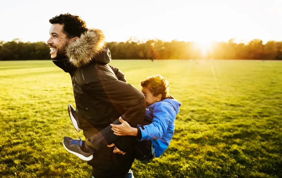 A single dad out with his son at the park, giving him a piggy back ride.