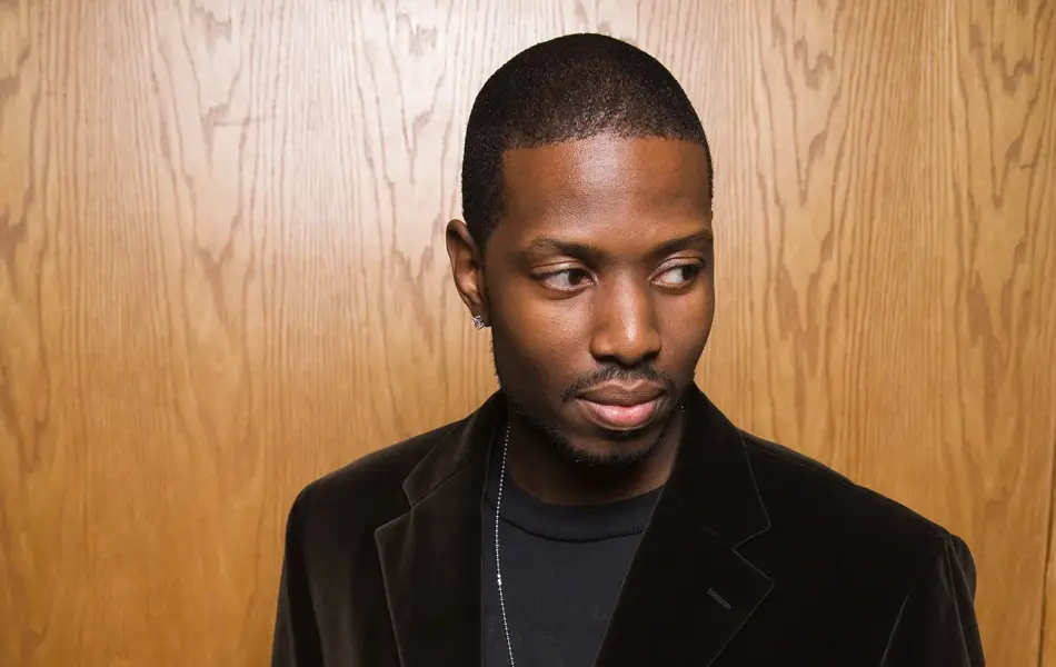 Mann med sort fløyelsjakke poserer foran trevegg 