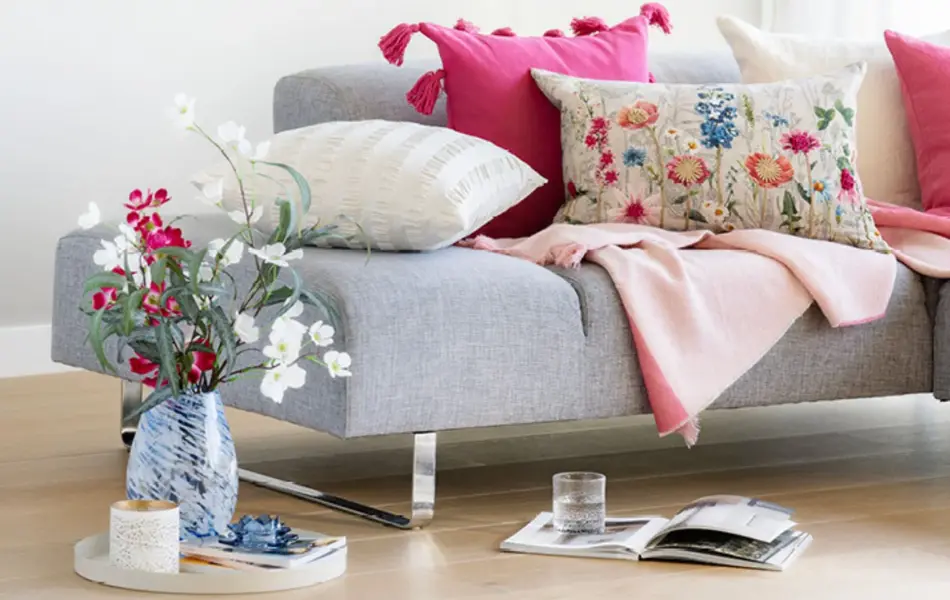 Grå sofa med fargerike puter, vase med blomster og en bok på gulvet