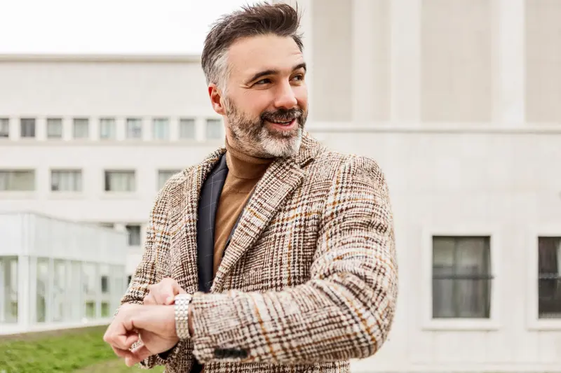 Mann med mørk og lys brun tweedjakke med ruter holder opp armen med klokke og smiler 