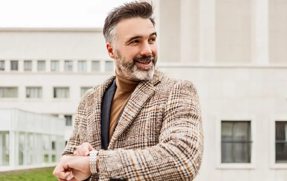 Mann med mørk og lys brun tweedjakke med ruter holder opp armen med klokke og smiler 