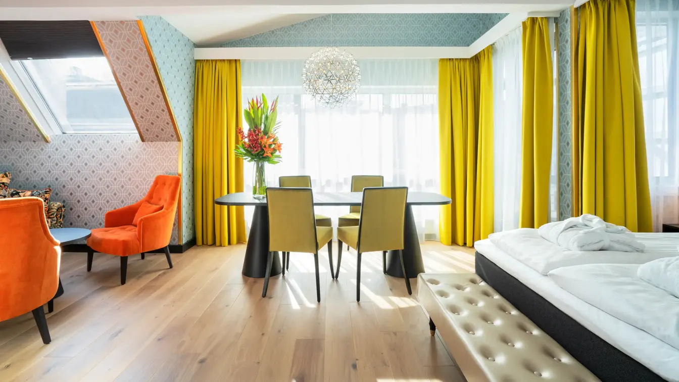 Sittegruppe med to enkeltstoler, stort bord med sitteplasser og store vinduer med store gule gardiner, kanten av dobbeltseng på business room på Thon Hotel Cecil i Oslo sentrum