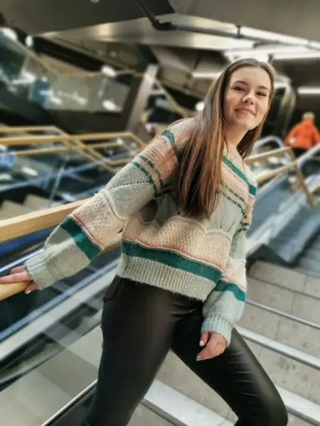Jente står i trapp på kjøpesenter, smiler til fotografen. Har på seg svarte skinnbukser og en strikkegenser med striper i fargene, aqua, grønn, dus rosa og kremhvit. 