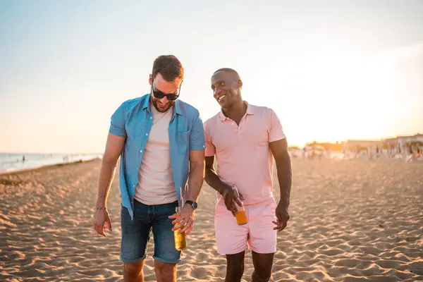 To menn går ved siden av hverandre på stranda. Den ene ser på den andre og smiler. Den ene mannen er iført en rosa shorts og en rosa pikéskjorte. Den andre har denimshorts, rosa t-skjorte og åpen denimskjorte over. Begge har en flaske i hånden.