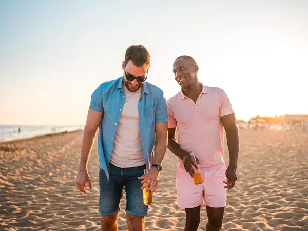 To menn går ved siden av hverandre på stranda. Den ene ser på den andre og smiler. Den ene mannen er iført en rosa shorts og en rosa pikéskjorte. Den andre har denimshorts, rosa t-skjorte og åpen denimskjorte over. Begge har en flaske i hånden.
