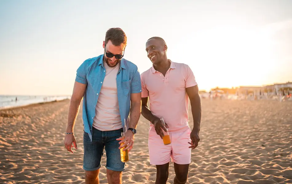 To menn går ved siden av hverandre på stranda. Den ene ser på den andre og smiler. Den ene mannen er iført en rosa shorts og en rosa pikéskjorte. Den andre har denimshorts, rosa t-skjorte og åpen denimskjorte over. Begge har en flaske i hånden.