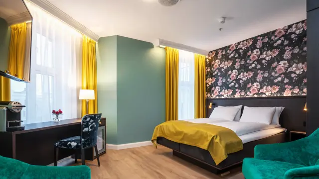 Mørk tapet med store lyserosa roser. Vindu med gule gardiner fra tak til gulv. Dobbeltseng med gult sengeteppe. Mørkt skrivebord med kaffemaskin og mønstrete stol. I superior rom på Thon Hotel Høyers i Skien.