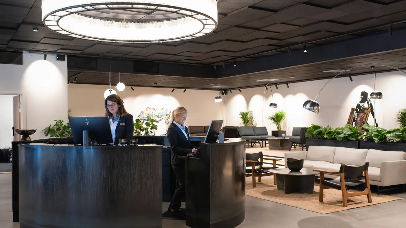 Rund mørk resepsjonsdisk med stor rund vegghengt lampe over og to resepsjonister. Lyse vegger og gulv og sort tak i resepsjonen på Thon Hotel Norge i Kristiansand. 