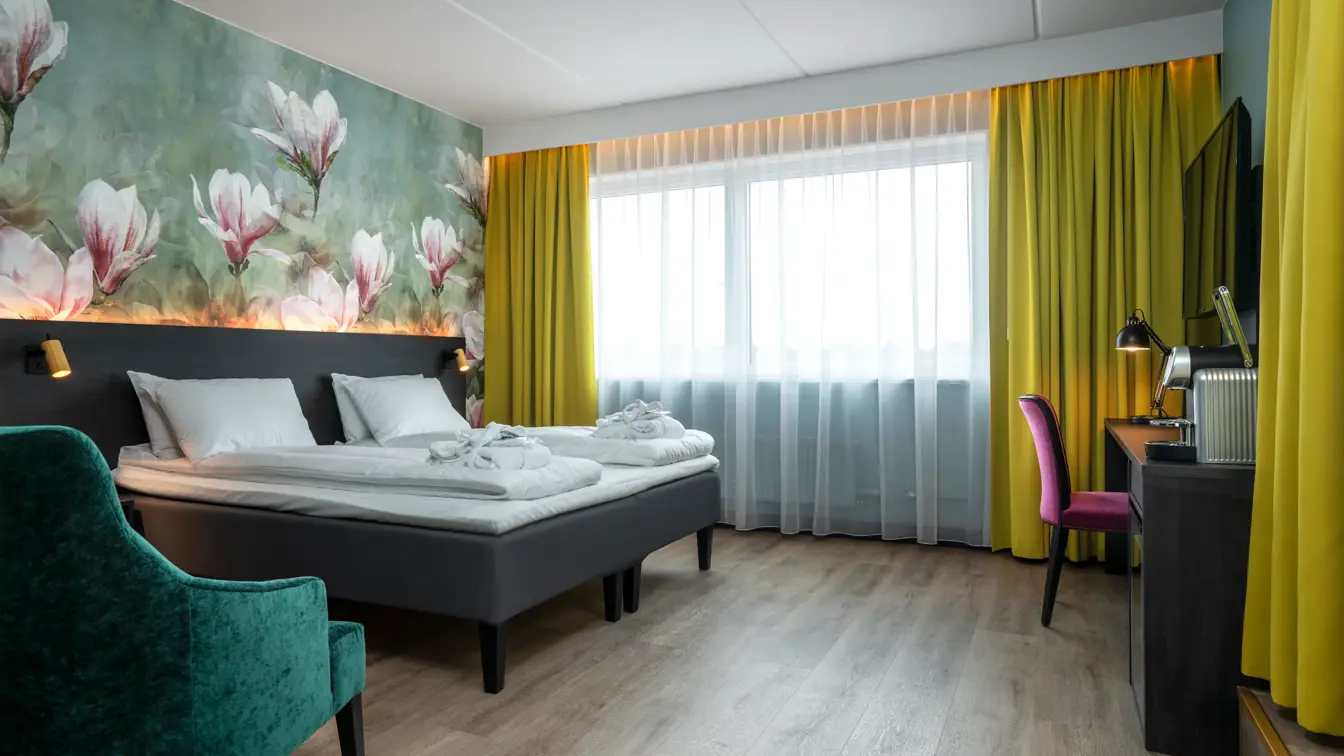 Blomstertapetvegg, dobbeltseng, vindu, gule gardiner, skrivebord, TV, rosa stol, grønn lenestol