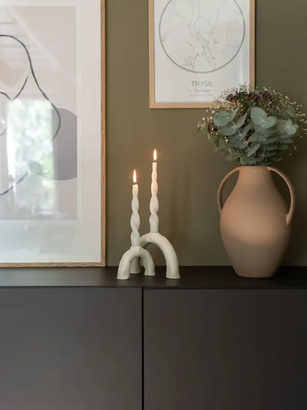 Hvite lysestaker med brennende lys og en grønnplante som står på en kommode