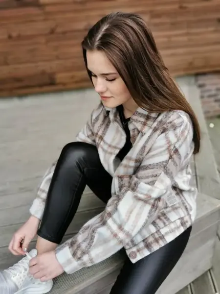 Jente sitter å knyter skoene ute på en benk. har på seg hvite sneakers, svart skinntights og en skjortejakke med ruter i dus brun, beige og rosa. 