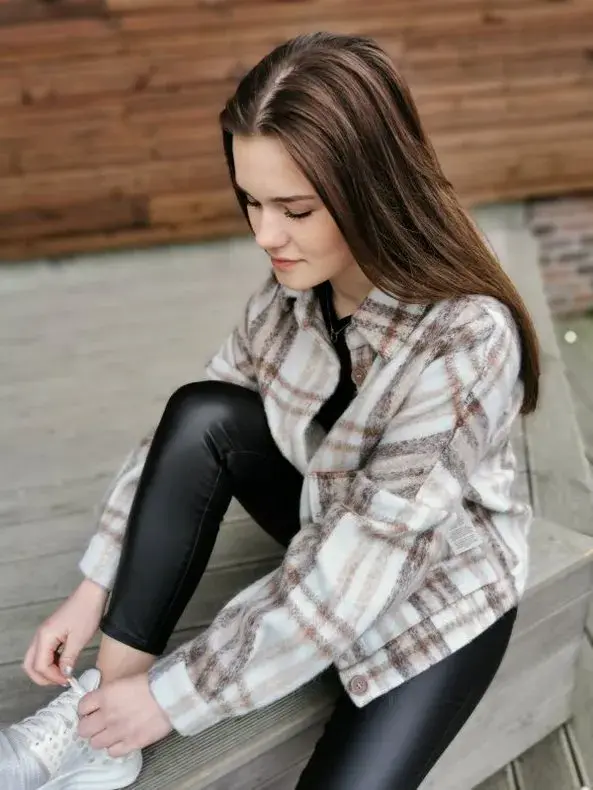 Jente sitter å knyter skoene ute på en benk. har på seg hvite sneakers, svart skinntights og en skjortejakke med ruter i dus brun, beige og rosa. 