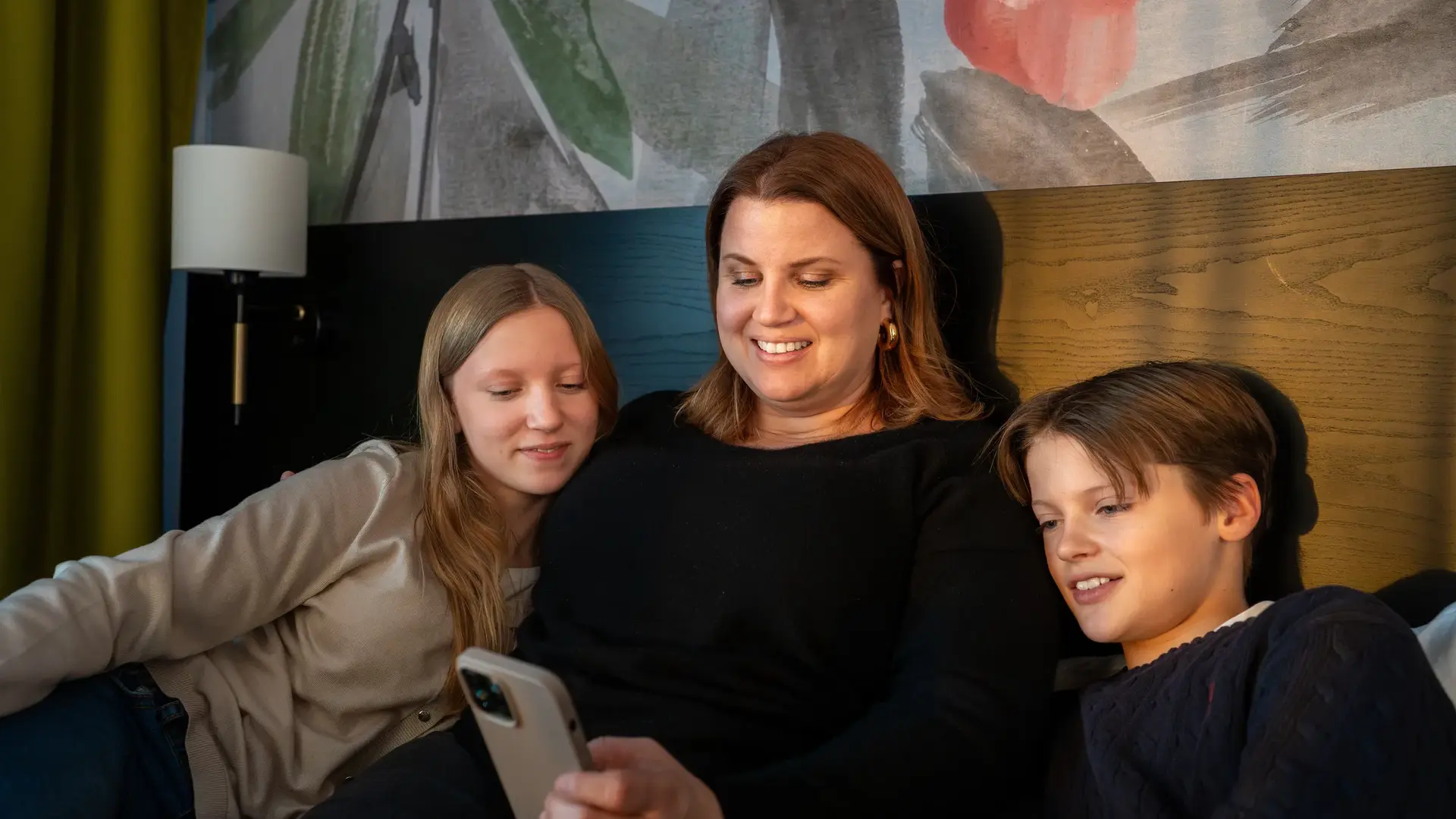 Kvinne og to barn sitter i en hotellseng og ser på en mobiltelefon.