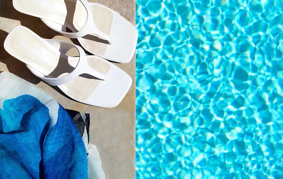 Nærbilde av hvite skinnflippflopper med hæl stående på bassengkant med turkist vann rundt 