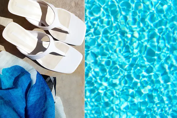 Nærbilde av hvite skinnflippflopper med hæl stående på bassengkant med turkist vann rundt 