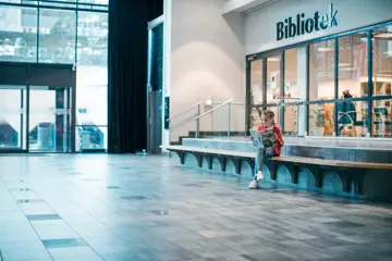 Jente leser en bok utenfor biblioteket ved inngangspartiet til Vestkanten Storsenter
