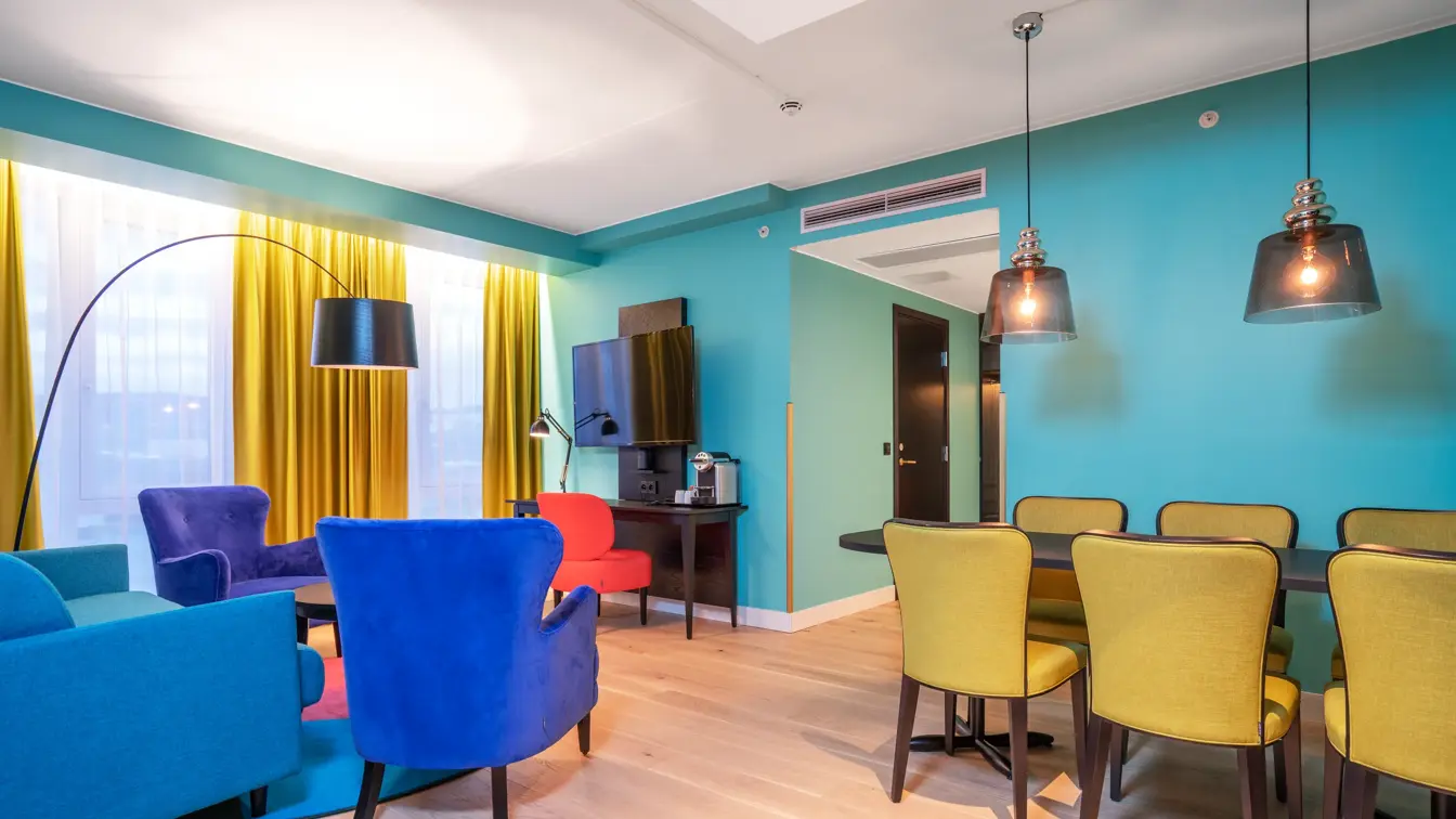 Turkis tapet, gule gardiner, sittegruppe med turkis sofa og blå stoler, spisebord med gule stoler og mørkt bord i junior suite på Thon Hotel Storo i Oslo.