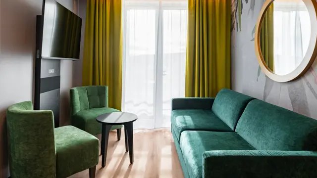 Stue i junior suite med grønne møbler, gule gardiner, tapet med mønster og parkettgulv.