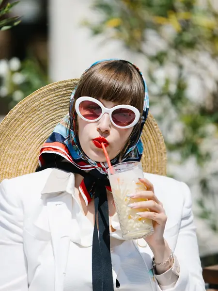 Halvportrett av dame med store hvite solbriller, rød leppestift, hvit jakke og silketørkle på hodet som drikker milkshake 
