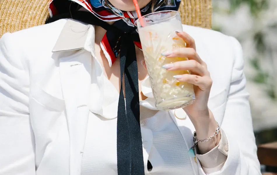 Halvportrett av dame med store hvite solbriller, rød leppestift, hvit jakke og silketørkle på hodet som drikker milkshake 