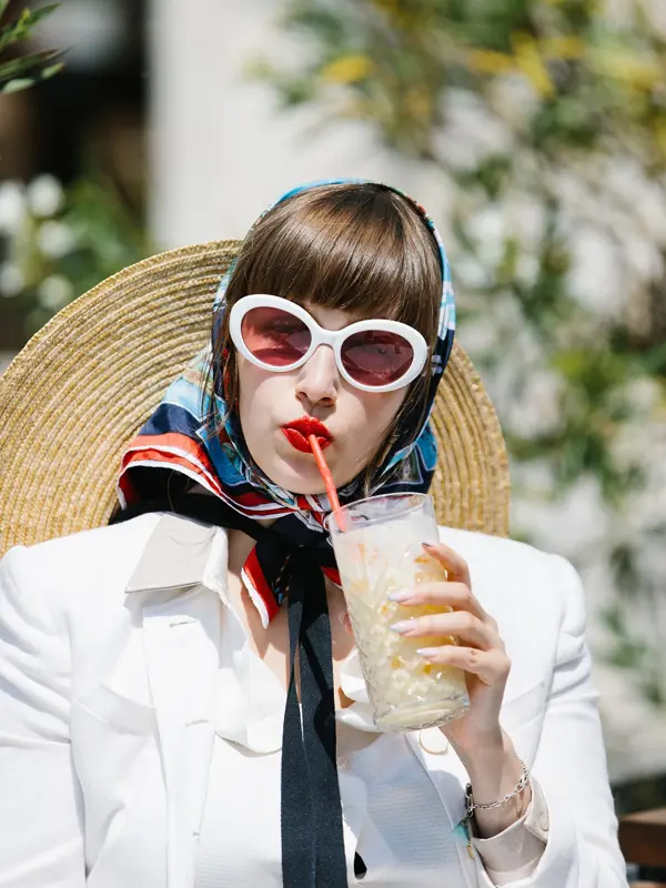 Halvportrett av dame med store hvite solbriller, rød leppestift, hvit jakke og silketørkle på hodet som drikker milkshake 