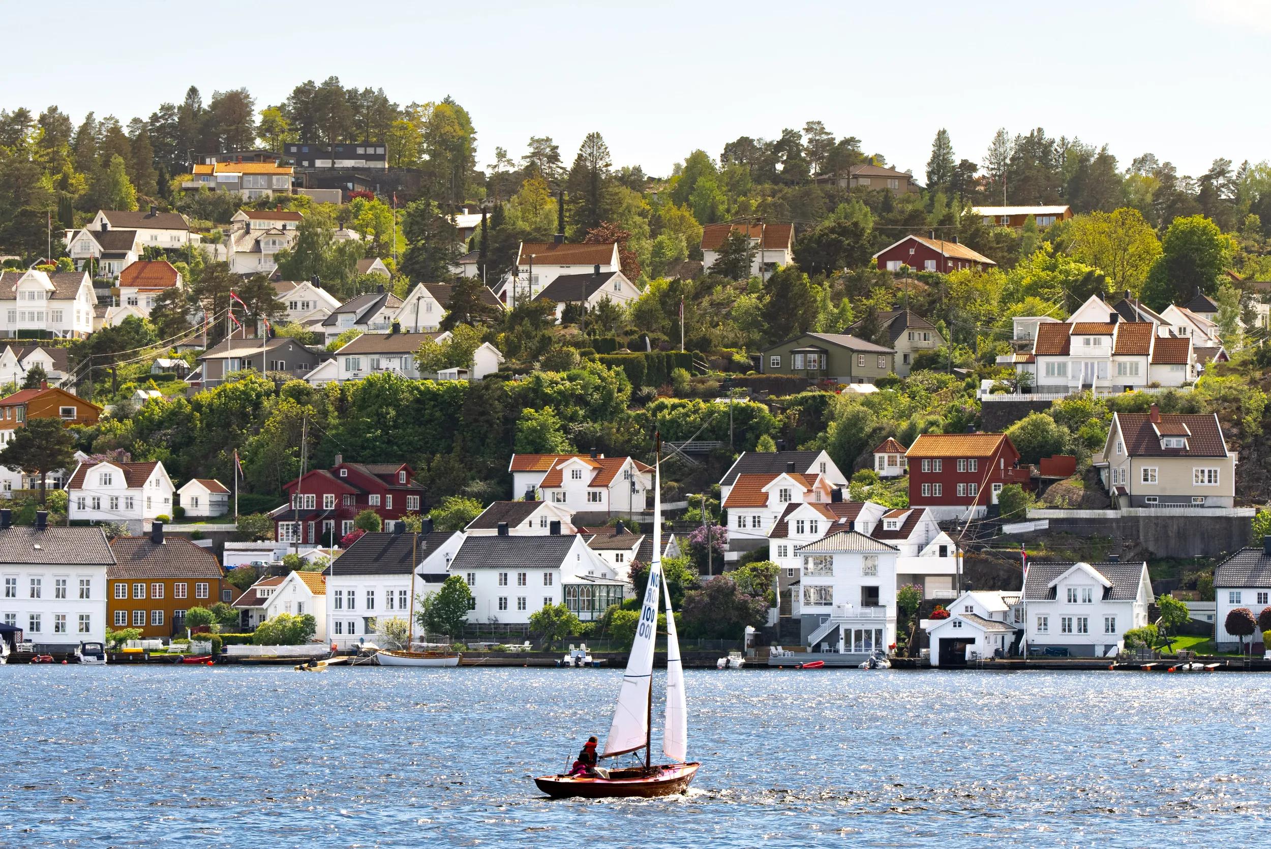 Seilb&aring;t med utsl&aring;tt seil foran sm&aring;hus bebyggelse i Arendal
