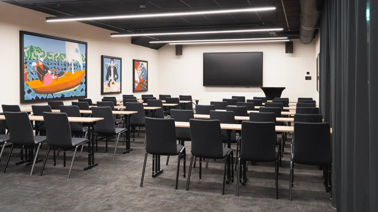 Møterom med klasseroms oppsett sett bakfra i lokalet. Svarte stoler og lyse trebord. TV skjerm foran.