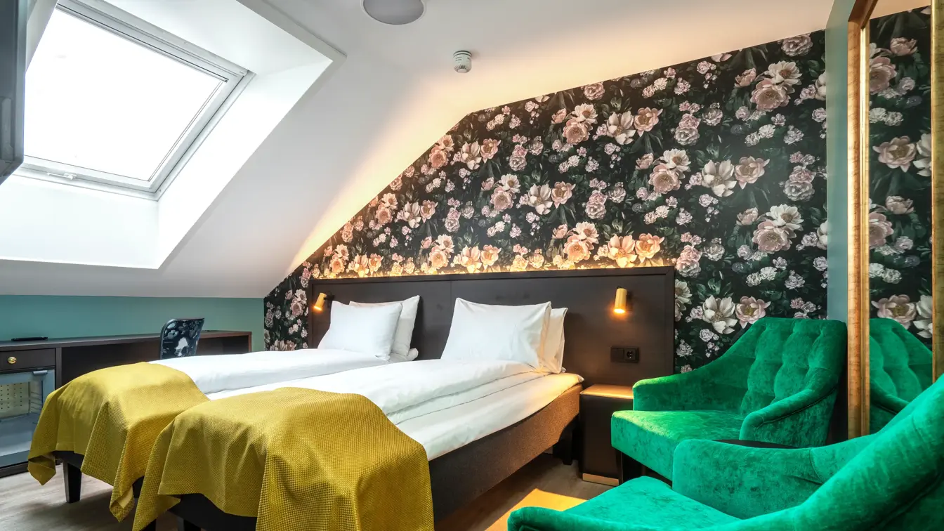 Skråtak med vindu. Mørk tapet med store lyserosa roser. To enkeltsenger med gule sengetepper. To grønne lenestoler. I standard twin rom på Thon Hotel Høyers i Skien.