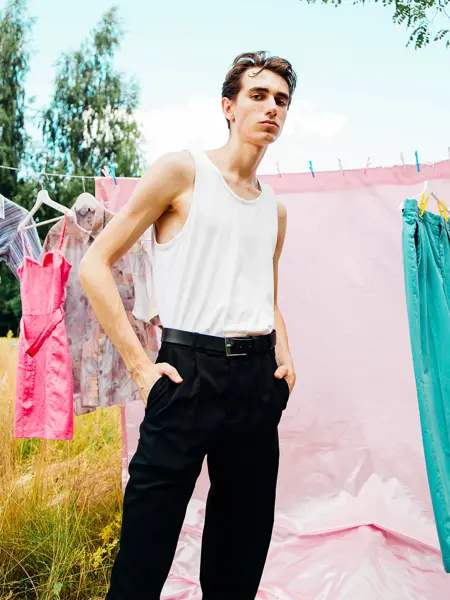 En mann poserer foran en tørkesnor med rosa klær til tørk. Mannen har hvit singlet og svarte bukser. Han har hendene i lommene, og ser rett i kamera.