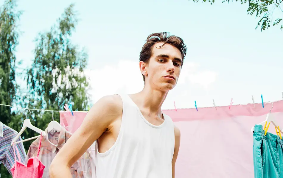 En mann poserer foran en tørkesnor med rosa klær til tørk. Mannen har hvit singlet og svarte bukser. Han har hendene i lommene, og ser rett i kamera.