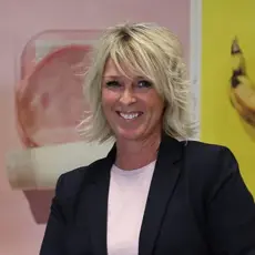 Smilende dame med blondt hår og sort dressjakke ser mot kamera, vegg med fargerike bilder bak.