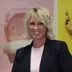 Smilende dame med blondt hår og sort dressjakke ser mot kamera, vegg med fargerike bilder bak.